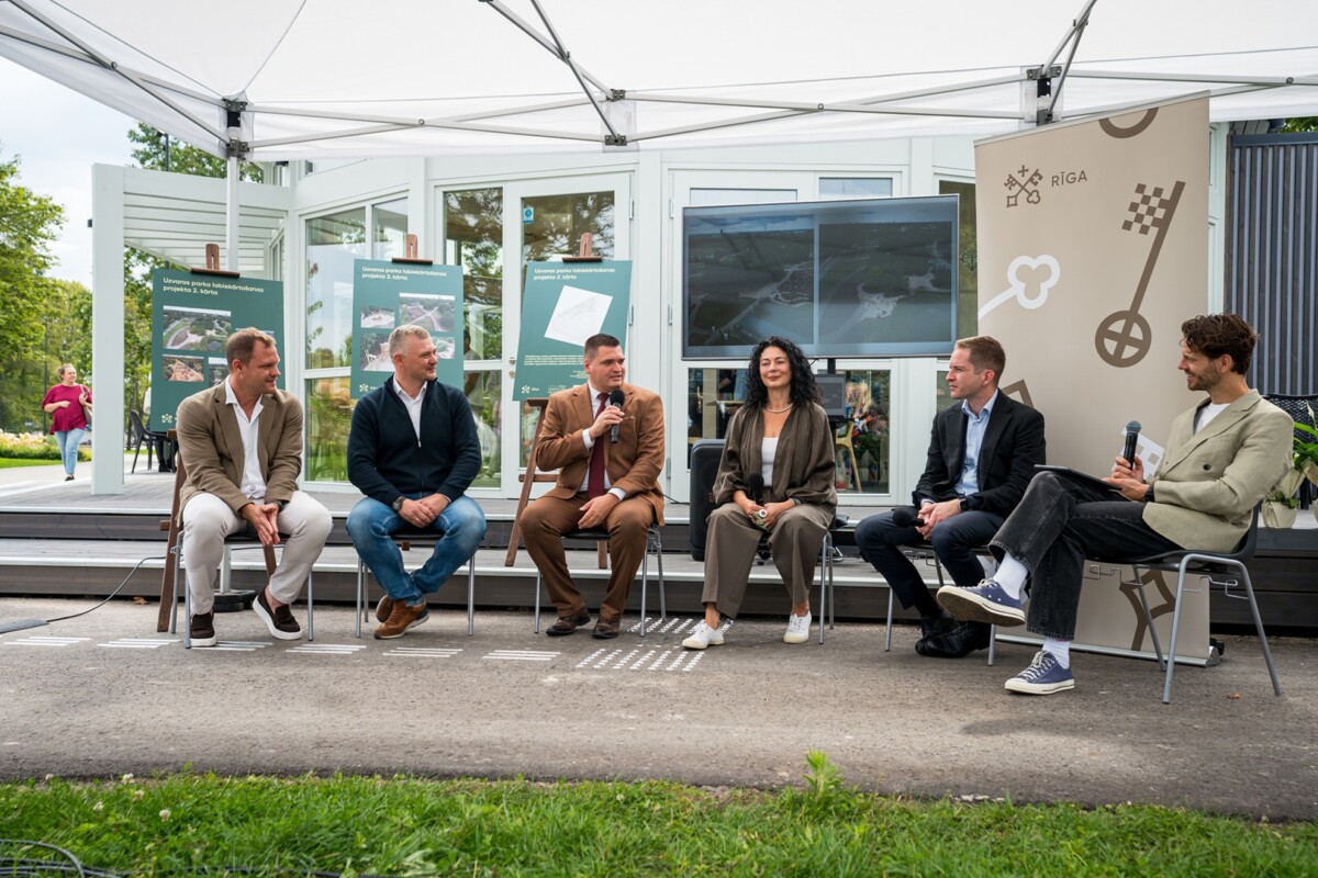 Seši cilvēki sēž uz krēsliem zem baltas nojumes āra pasākumā; fonā redzami stendi ar uzrakstiem 'UZVARAS PARKS' un 'RIGA', apkārt redzama zaļa vide