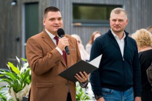 Divi cilvēki stāv blakus; viens tur mikrofonu un mapi, iespējams, uzrunā klātesošos, otrs stāv blakus; fonā redzami augi un citi cilvēki, norisinās āra pasākums