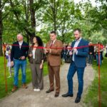Āra lentes griešanas ceremonija ar četriem cilvēkiem, kas tur šķēres pie sarkanās lentes; fonā skatītāji un zaļa parka vide ar kokiem un zāli