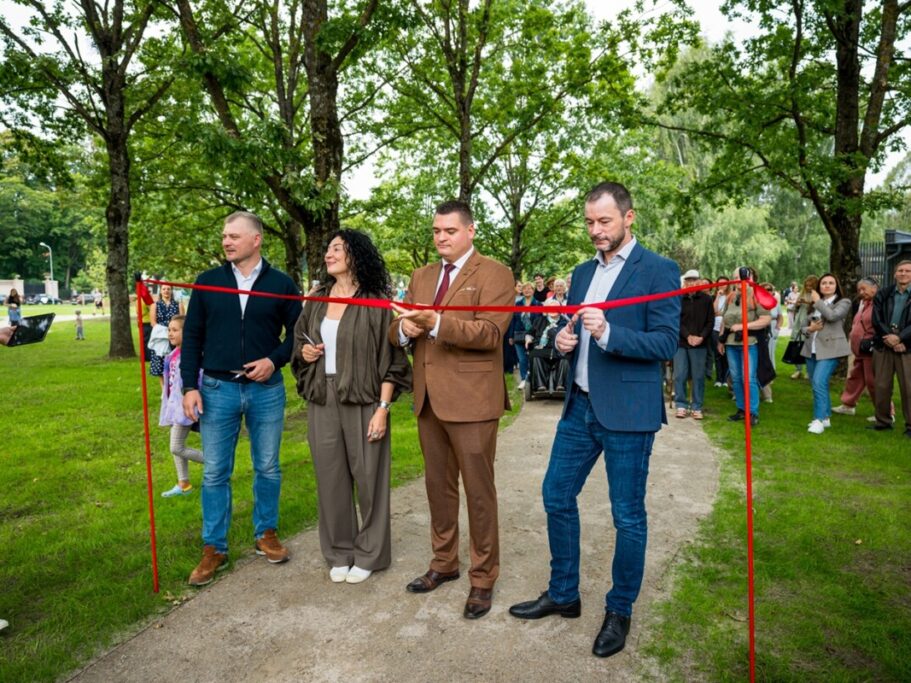 Āra lentes griešanas ceremonija ar četriem cilvēkiem, kas tur šķēres pie sarkanās lentes; fonā skatītāji un zaļa parka vide ar kokiem un zāli