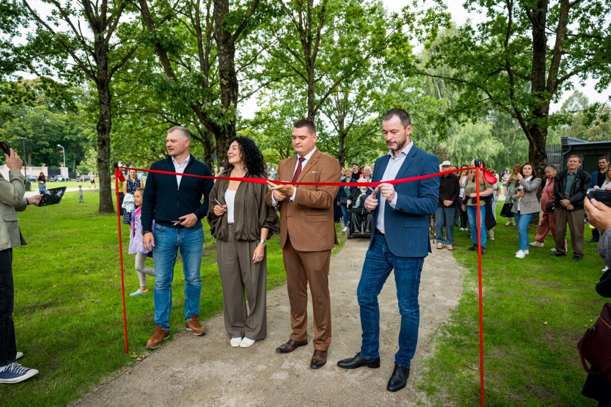 Āra lentes griešanas ceremonija ar četriem cilvēkiem, kas tur šķēres pie sarkanās lentes; fonā skatītāji un zaļa parka vide ar kokiem un zāli