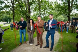 Āra pasākums ar sarkanās lentes griešanas ceremoniju; četri cilvēki tur lenti, viens centrā to pārgriež ar šķērēm, fonā redzama cilvēku grupa un zaļa parka vide