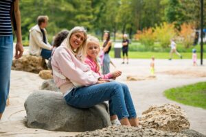 Cilvēki parkā; priekšplānā divas personas sēž uz liela akmens, tērptas rozā un zilā apģērbā, fonā redzami koki, celiņi un citi cilvēki pastaigājas vai stāv
