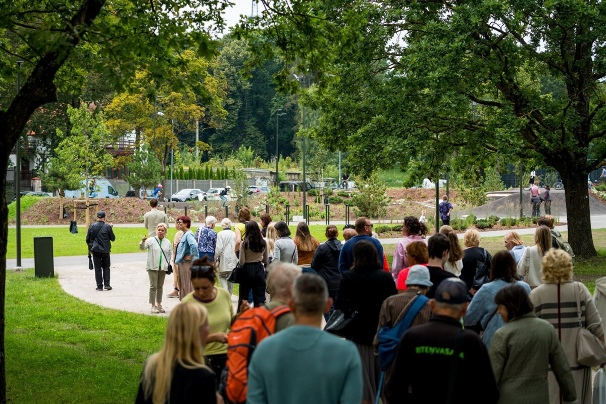 Cilvēku grupa pastaigājas pa parku ar zaļu zāli, kokiem un celiņiem; daži nēsā mugursomas un jakas, fonā redzami koki un stāvošas automašīnas
