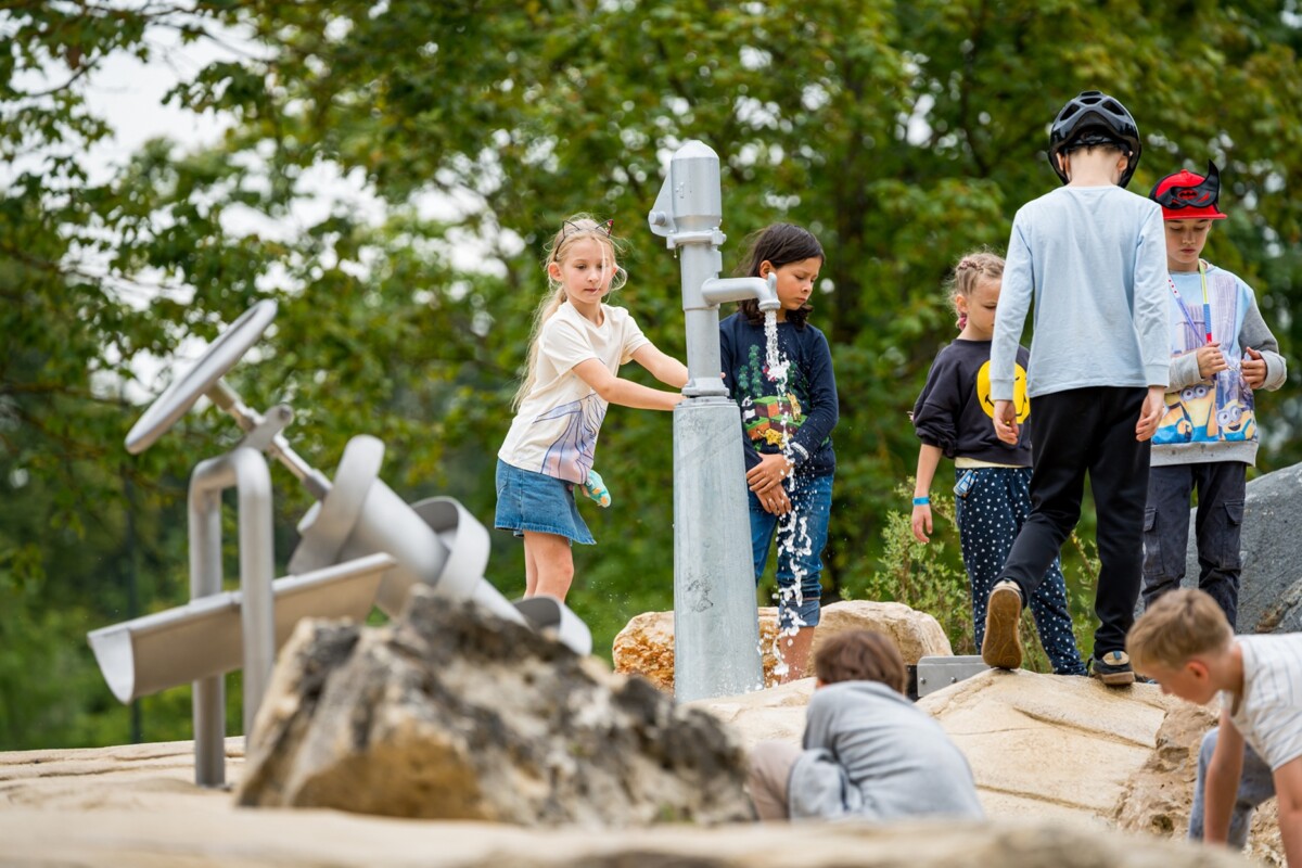 Bērni rotaļājas dabas vidē pie ūdens sūkņa; viens bērns darbojas ar sūkni, citi vēro un piedalās dažādās aktivitātēs, fonā koki un akmeņi