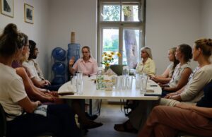 Grupa cilvēku sēž ap taisnstūrveida galdu sapulču telpā; uz galda redzamas glāzes un uzkodas. Fonā logs ar skatu uz kokiem, ūdens aparāts ar pudelēm un sienas ar ierāmētām fotogrāfijām