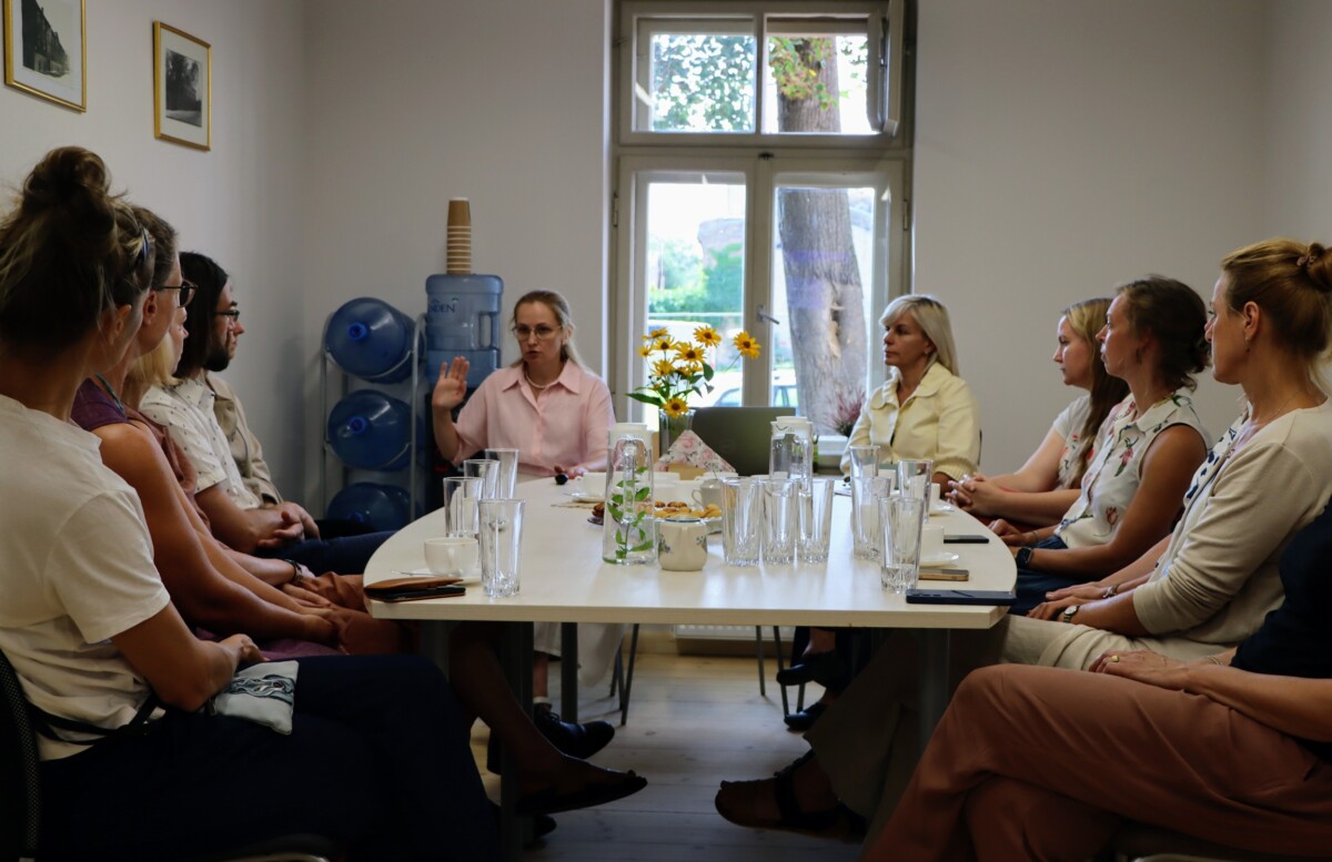 Grupa cilvēku sēž ap taisnstūrveida galdu sapulču telpā; uz galda redzamas glāzes un uzkodas. Fonā logs ar skatu uz kokiem, ūdens aparāts ar pudelēm un sienas ar ierāmētām fotogrāfijām