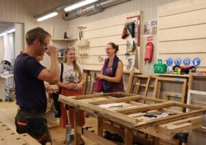 Kokapstrādes darbnīca ar vairākiem cilvēkiem ap darba galdu, uz kura redzami instrumenti, koka rāmji un papīri. Fonā pie sienām izvietoti darbarīki, drošības zīmes, skavas, zāģi.