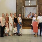 Grupa cilvēku stāv telpā pie veca koka loga ar slēģiem; daži atrodas loga rāmī, citi – ārpusē. Redzamas krāsainas auduma somas, fonā – vienkārša siena un koka konstrukcija