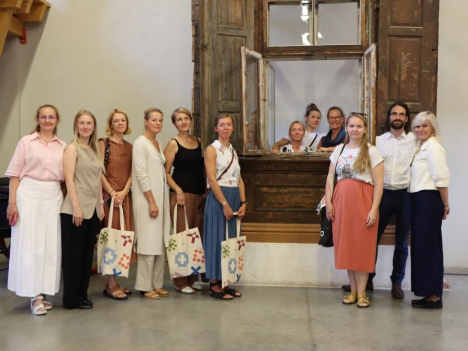 Grupa cilvēku stāv telpā pie veca koka loga ar slēģiem; daži atrodas loga rāmī, citi – ārpusē. Redzamas krāsainas auduma somas, fonā – vienkārša siena un koka konstrukcija