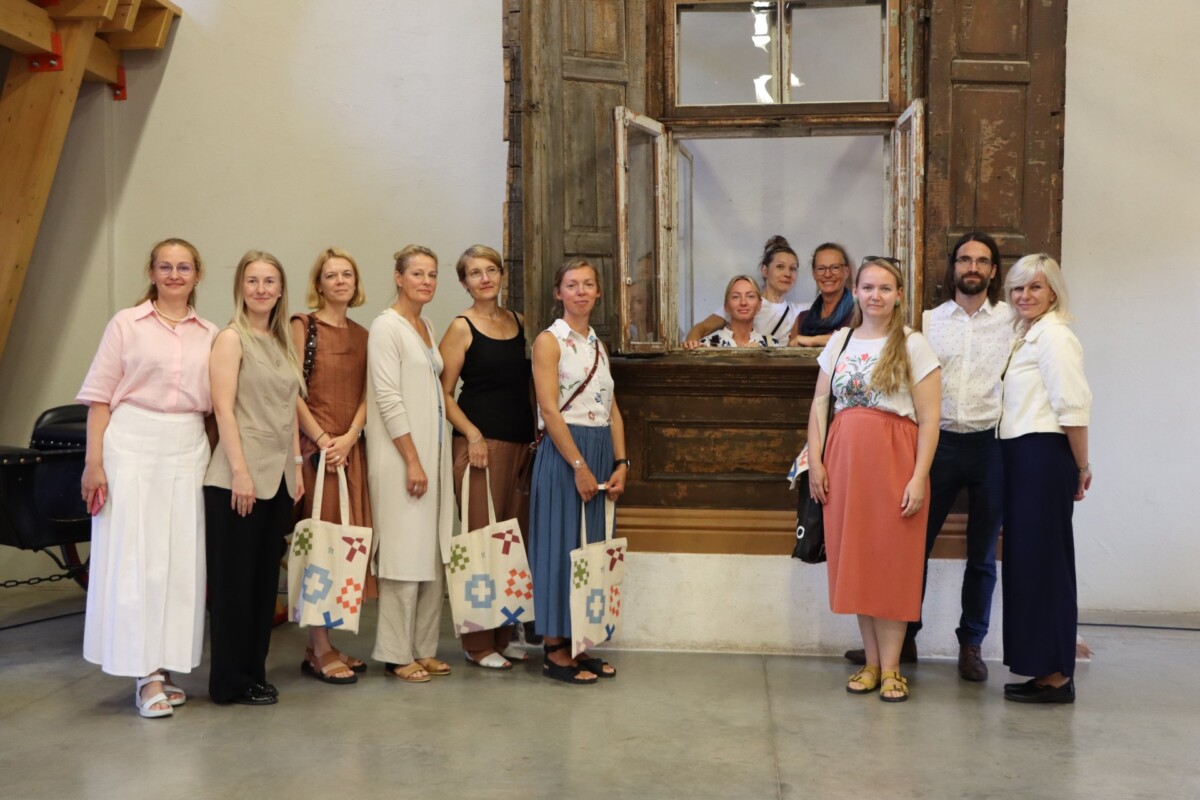 Grupa cilvēku stāv telpā pie veca koka loga ar slēģiem; daži atrodas loga rāmī, citi – ārpusē. Redzamas krāsainas auduma somas, fonā – vienkārša siena un koka konstrukcija