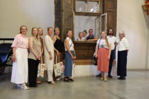 Grupa cilvēku stāv pie veca koka loga ar atvērtām slēģiem; daži atrodas loga iekšpusē un mijiedarbojas ar tiem, kas ir ārpusē. Redzamas somas ar dažādiem rakstiem, telpa izskatās kā vēsturiska vai kultūras vieta