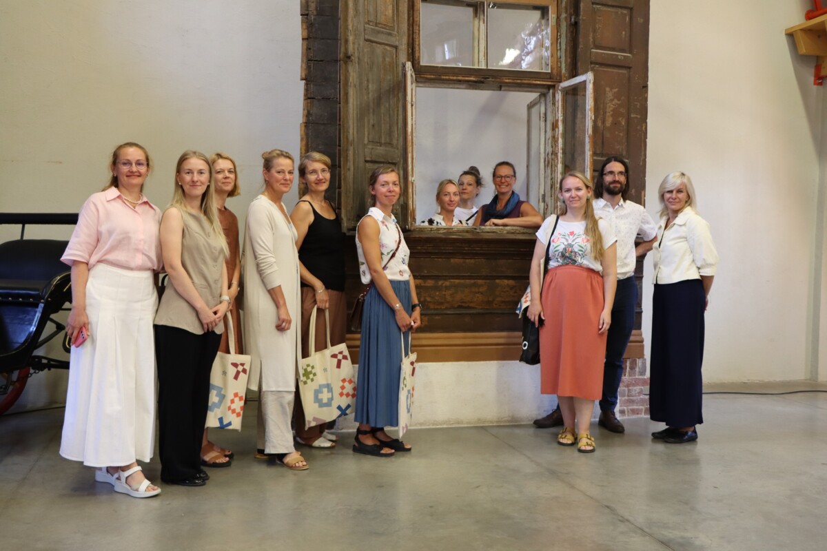 Grupa cilvēku stāv pie veca koka loga ar atvērtām slēģiem; daži atrodas loga iekšpusē un mijiedarbojas ar tiem, kas ir ārpusē. Redzamas somas ar dažādiem rakstiem, telpa izskatās kā vēsturiska vai kultūras vieta