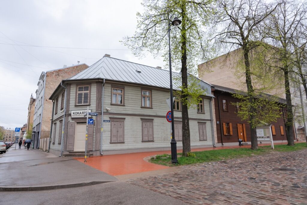 Koka ēkas ārpuse ar horizontālām dēlēm, vecām koka slēģiem pie loga un bruģētu celiņu priekšplānā