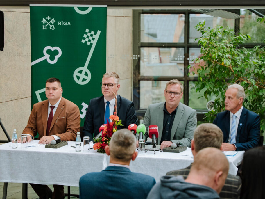 Preses konference ar četriem cilvēkiem pie galda, uz kura novietoti dažādu mediju mikrofoni