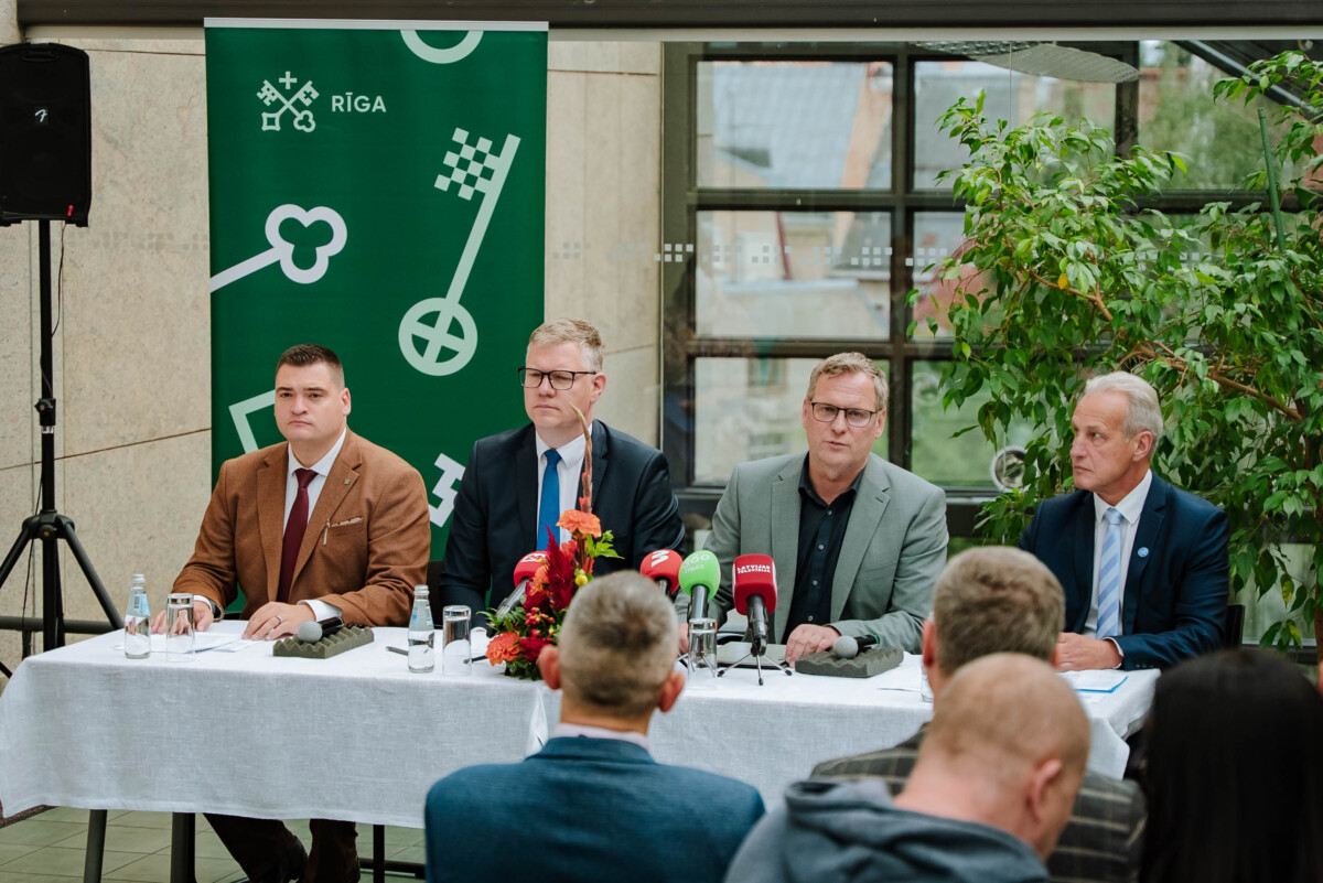 Preses konference ar četriem cilvēkiem pie galda, uz kura novietoti dažādu mediju mikrofoni