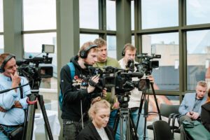 Cilvēku grupa telpā ar lieliem logiem darbojas ar videokamerām un ierakstu aprīkojumu; redzami statīvi, austiņas un skats uz ēkām fonā