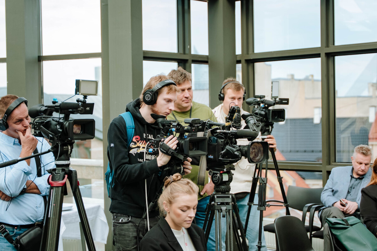 Cilvēku grupa telpā ar lieliem logiem darbojas ar videokamerām un ierakstu aprīkojumu; redzami statīvi, austiņas un skats uz ēkām fonā