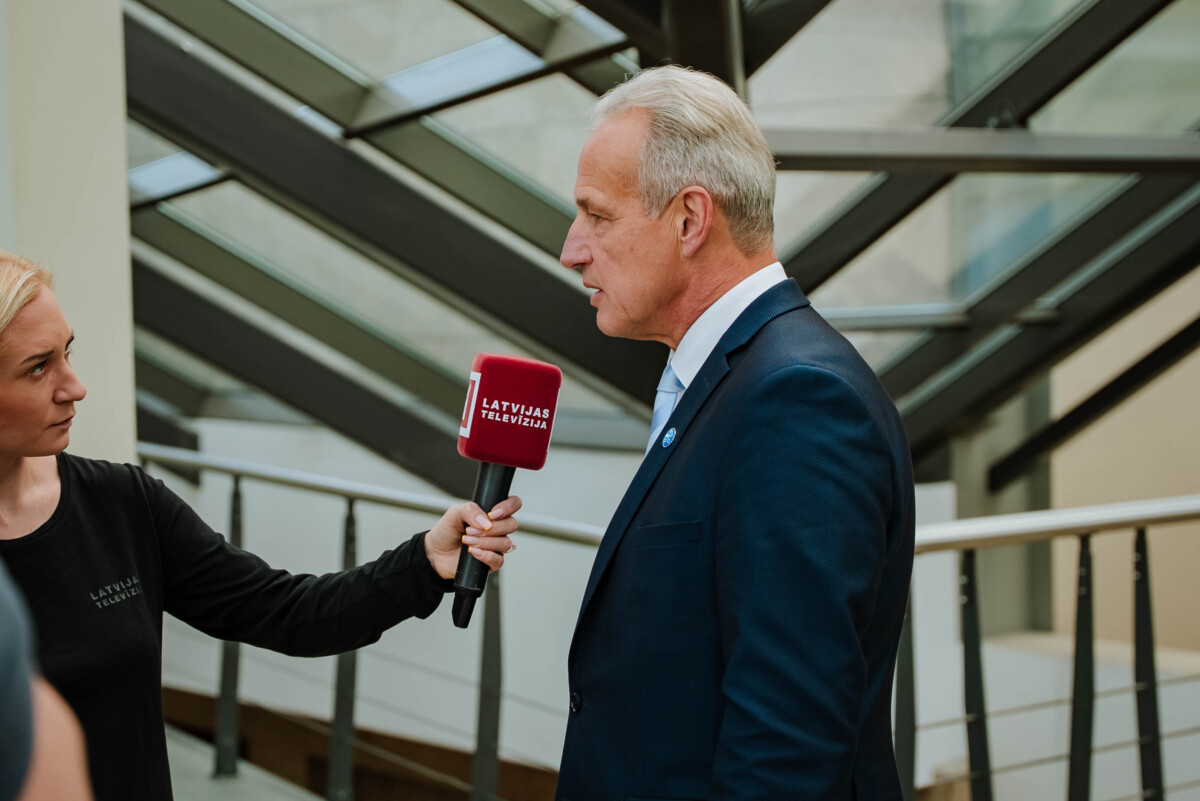 Žurnālists ar mikrofonu 'LATVIJAS TELEVĪZIJA' intervē personu pie mūsdienīgas stikla un metāla arhitektūras fona