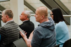 Telpā sēdoša cilvēku grupa; priekšplānā persona zilā džemperī tur viedtālruni