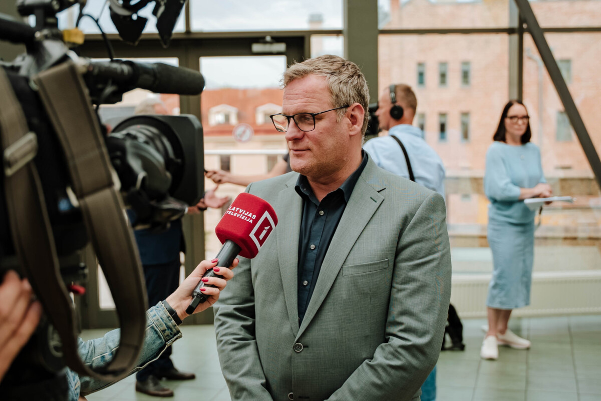 Notiek intervija, intervētājam rokā mikrofons ar 'LRT TELEVIZIJA' logo, fonā redzams operatora filmēšanas aprīkojums un divi cilvēki, no kuriem viens raksta uz klades