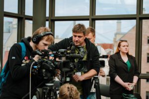 Grupa cilvēku strādā ar profesionālu video ieraksta aprīkojumu telpā ar lieliem logiem, fonā redzami pilsētas ēku skati