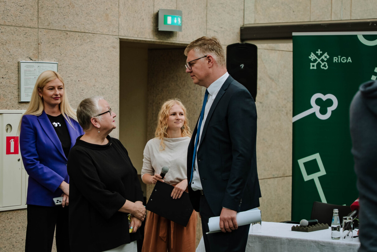 Četru cilvēku grupa sarunājas telpā; viens tur mikrofonu, otrs – dokumentus; fonā zaļš plakāts ar uzrakstu 'RĪGA