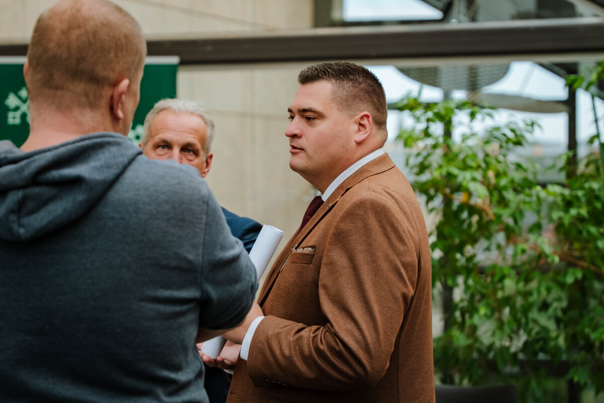 Trīs cilvēki sarunājas; fonā redzami augi un ēkas struktūra