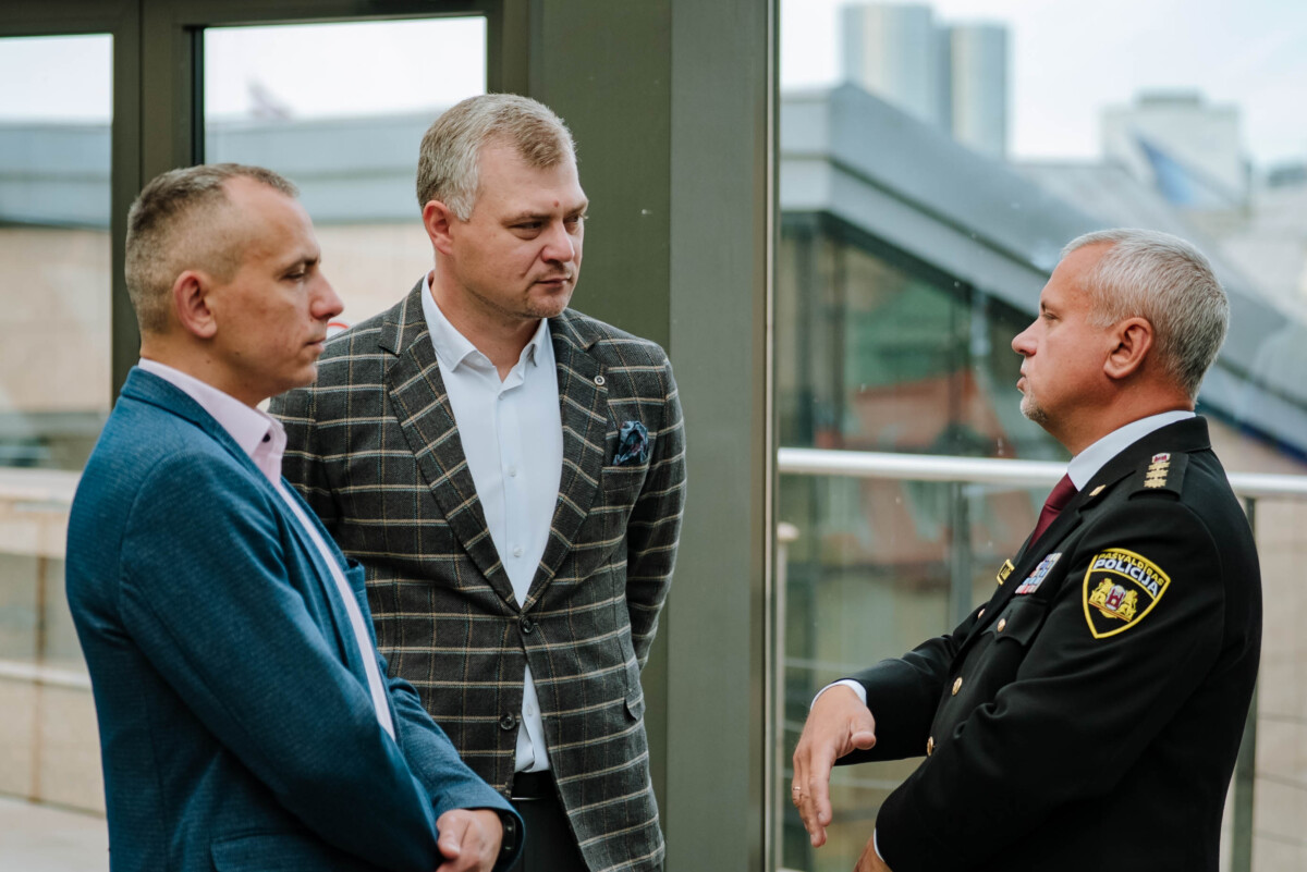 Trīs vīrieši sarunājas; divi tērpti uzvalkos, viens – policijas formā; fonā lieli logi ar skatu uz pilsētas ēkām