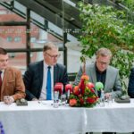 Preses konference ar četriem cilvēkiem pie galda, uz kura novietoti mikrofoni un ziedu kompozīcija; fonā lieli logi un zaļumi