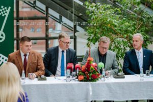 Preses konference ar četriem cilvēkiem pie galda, uz kura novietoti mikrofoni un ziedu kompozīcija; fonā lieli logi un zaļumi