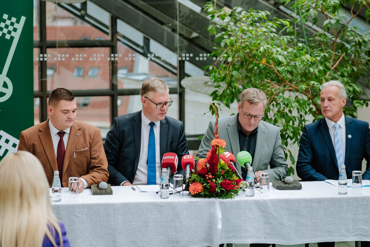 Preses konference ar četriem cilvēkiem pie galda, uz kura novietoti mikrofoni un ziedu kompozīcija; fonā lieli logi un zaļumi