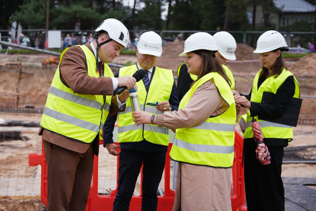 "Cilvēku grupa ar baltām aizsargķiverēm un dzeltenām atstarojošām vestēm būvlaukumā; viens cilvēks runā mikrofonā, cits tur metāla cilindru, fonā redzamas sarkanas barjeras un būvniecības aprīkojums