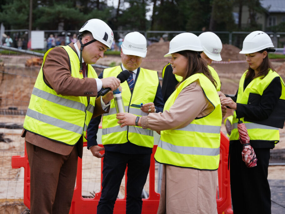 "Cilvēku grupa ar baltām aizsargķiverēm un dzeltenām atstarojošām vestēm būvlaukumā; viens cilvēks runā mikrofonā, cits tur metāla cilindru, fonā redzamas sarkanas barjeras un būvniecības aprīkojums