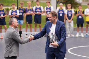 Divi vīrieši sarokojas basketbola laukumā; viens tur mikrofonu, otrs – iesaiņotu dāvanu. Fonā redzami jaunieši sporta formās, kas stāv rindā