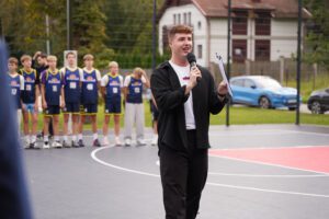 Persona ar mikrofonu un kladi stāv uz āra basketbola laukuma; fonā redzama sporta formās tērpta cilvēku grupa, ēkas un stāvošas automašīnas