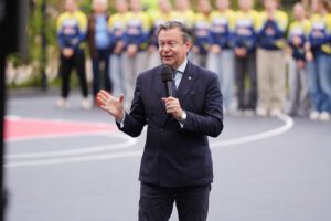 Persona uzvalkā runā mikrofonā ar paceltu roku āra sporta laukumā; fonā redzama cilvēku grupa vienādās zilās un dzeltenās formās