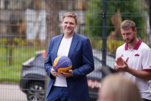 Divi cilvēki stāv ārā pie žoga; viens tērpts zilā uzvalkā un tur basketbola bumbu, otrs – baltā kreklā ar bordo akcentiem, šķietami aplaudē