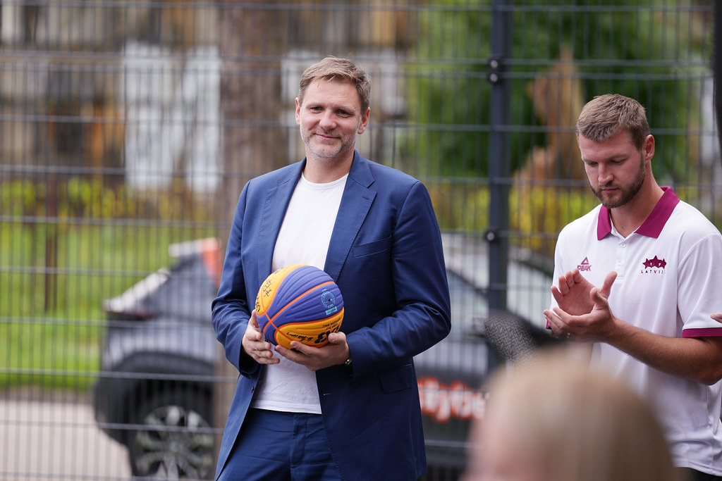 Divi cilvēki stāv ārā pie žoga; viens tērpts zilā uzvalkā un tur basketbola bumbu, otrs – baltā kreklā ar bordo akcentiem, šķietami aplaudē