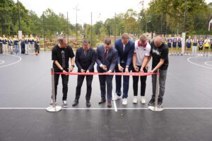 Seši cilvēki stāv rindā un pārgriež sarkanu lenti āra pasākumā, kas, šķiet, ir jauna sporta laukuma atklāšana; fonā redzami skatītāji un koki