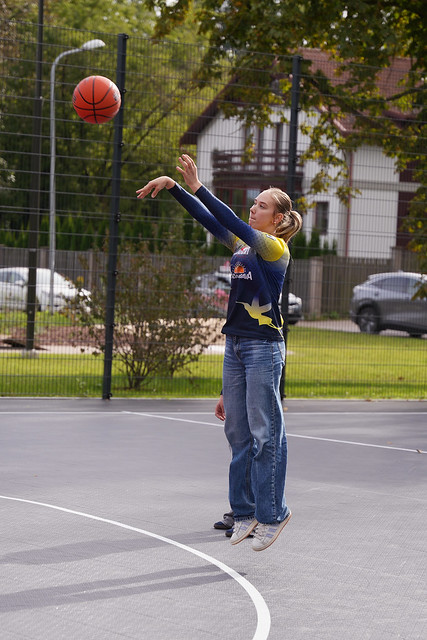 Persona met basketbola bumbu uz āra laukuma; bumba redzama gaisā, fonā – žogs, zaļumi un stāvošas automašīnas