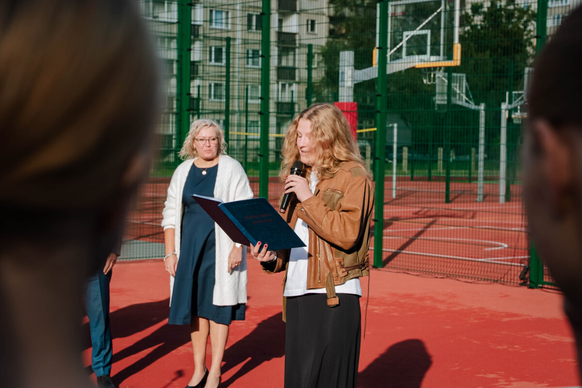 Cilvēku grupa stāv uz sarkana āra sporta laukuma, iespējams, pasākuma vai ceremonijas laikā; viens cilvēks runā mikrofonā un lasa no zilas mapes