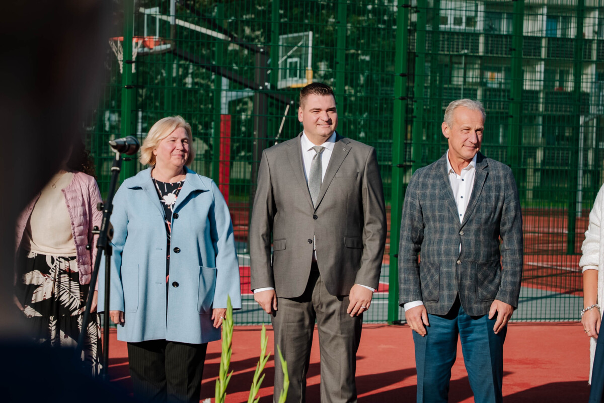Trīs cilvēki stāv uz sarkana sporta laukuma; fonā zaļš žogs un basketbola grozs; viens tērpts gaiši zilā mētelī, otrs pelēkā uzvalkā, trešais rūtainā žaketē
