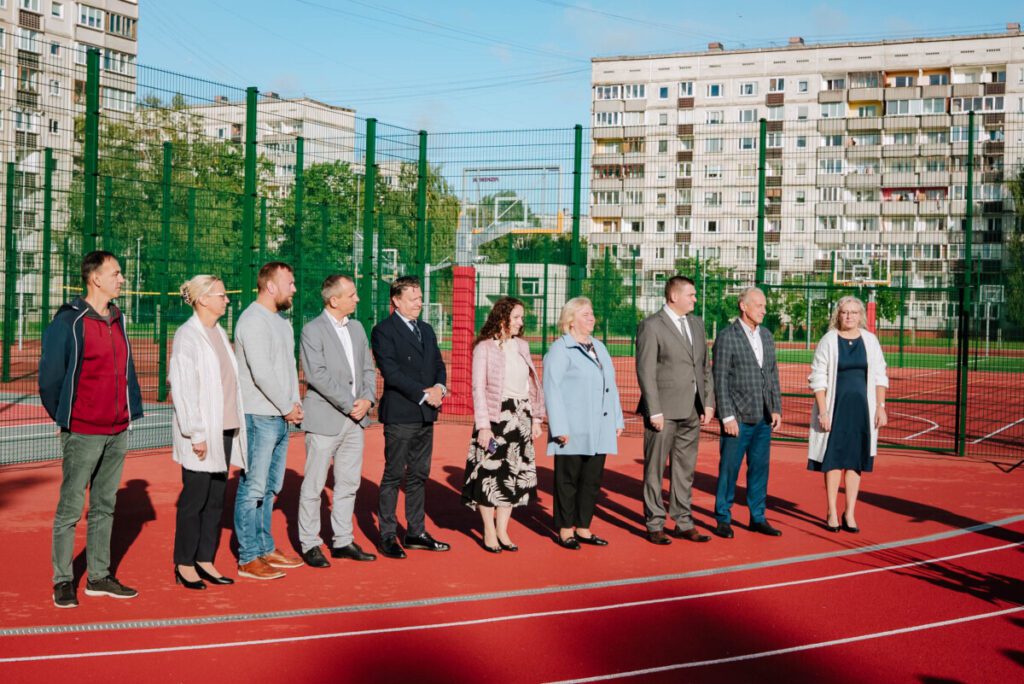 Deviņi cilvēki stāv rindā uz āra sporta laukuma; fonā zaļš žogs, basketbola grozi un daudzdzīvokļu ēkas
