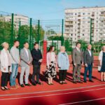 Deviņi cilvēki stāv rindā uz āra sporta laukuma; fonā zaļš žogs, basketbola grozi un daudzdzīvokļu ēkas
