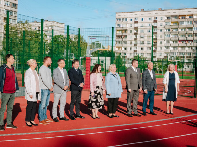 Deviņi cilvēki stāv rindā uz āra sporta laukuma; fonā zaļš žogs, basketbola grozi un daudzdzīvokļu ēkas