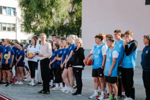 Cilvēku grupa stāv uz āra sporta laukuma; daļa tērpta zilās kreklos un tur rokās volejbola bumbas, otra daļa – gaiši zilās formās ar numuriem un basketbola bumbu