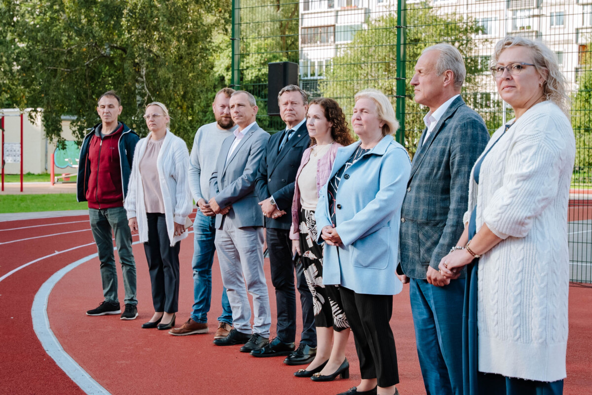 Deviņi cilvēki stāv rindā uz āra skrejceļa, tērpti formālā un pusformālā apģērbā; fonā redzami koki, žogs un ēkas daļa
