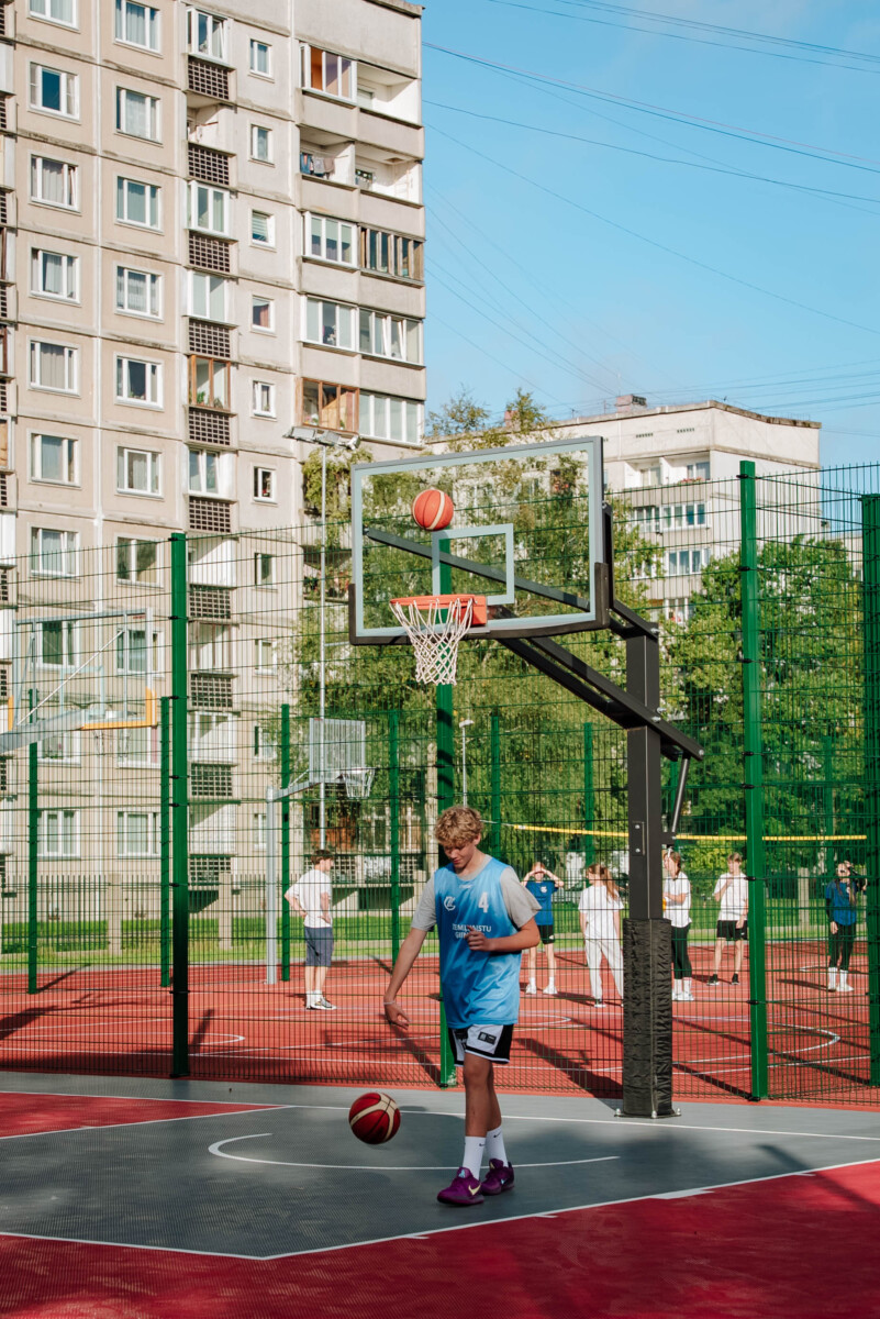 Āra basketbola laukums; jaunietis zilā kreklā un baltos šortos stāv pie groza ar bumbu rokās, cita bumba lido cauri grozam; fonā citi cilvēki un daudzdzīvokļu ēka; skaidrs dienas laiks