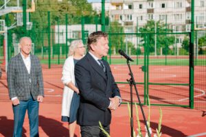 Trīs cilvēki stāv uz sarkana āra sporta laukuma; viens runā mikrofonā, pārējie divi stāv aiz viņa; fonā zaļš žogs, notiek pasākums vai ceremonija